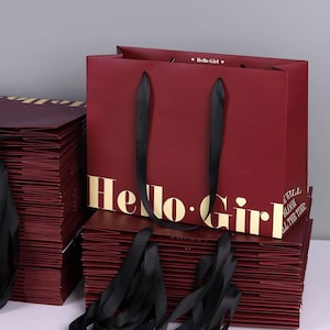 May include: Burgundy shopping bags with black ribbon handles. The bags have gold lettering that reads "Hello Girl" and "Will Bloom All The Time". A stack of bags is visible.