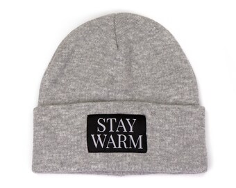 May include: A gray beanie hat with a black rectangular patch that reads "STAY WARM" in white capital letters. The hat is made of a soft, knitted material and is designed to keep the wearer warm in cold weather.