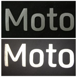 May include: A close-up of the word "Moto" in white reflective lettering on a black fabric background. The lettering is slightly raised and has a textured surface.
