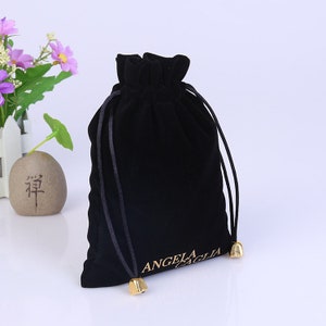 Puede incluir: Bolsa de cordón de terciopelo negro con cordón dorado y cierres de metal dorado. La bolsa tiene el texto "ANGELA ZAGLIA" impreso en dorado en la parte delantera.