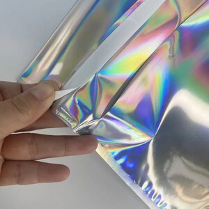 Puede incluir: Primer plano de un material de embalaje hologr&aacute;fico iridiscente. El material est&aacute; doblado por la mitad, revelando un respaldo plateado.