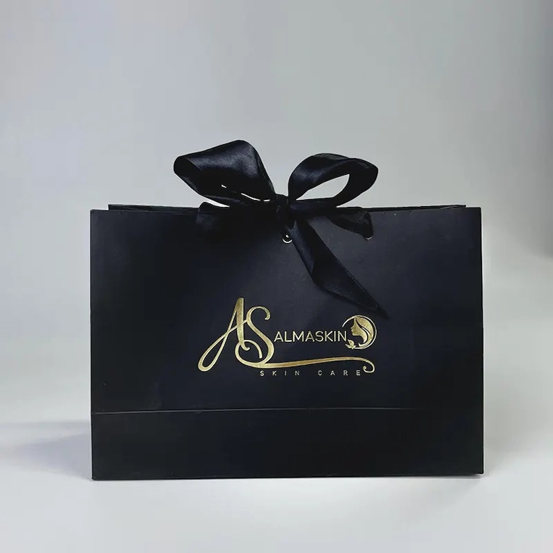 May include: Black gift bag with a satin ribbon bow. The bag features gold lettering that reads "AS ALMASKIN SKIN CARE" with a logo of a woman's face. The bag is a square shape and is likely used for packaging skincare products.