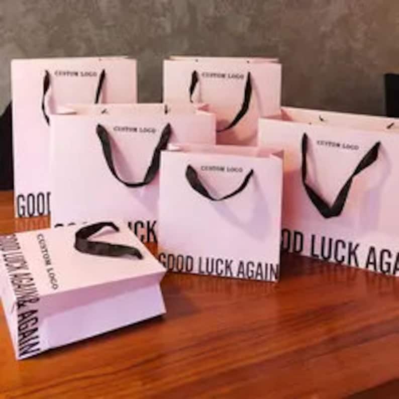 May include: Pink shopping bags with black handles are arranged on a wooden surface. The bags have the text "GOOD LUCK AGAIN" and "CUSTOM LOGO" printed on them. The bags are of varying sizes.