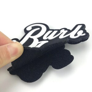 May include: A black and white rubber patch with the word "Burb" in white lettering. The patch has a hook and loop backing.