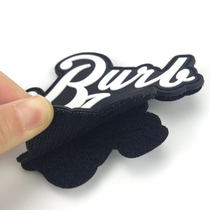 May include: A black and white rubber patch with the word "Burb" in white lettering. The patch has a hook and loop backing.