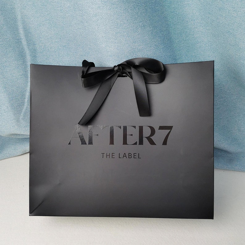 May include: A black gift bag with a satin ribbon bow at the top. The bag has the words "AFTER 7" and "THE LABEL" printed on the front in a dark gray color. The bag is sitting on a white surface with a light blue background.