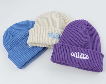 May include: Three ribbed knit beanies in blue, cream, and purple. Each beanie features the word "DAIZEE" embroidered on the folded brim. The beanies are displayed on a white background.