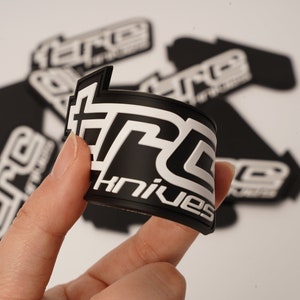 May include: Black and white TRC Knives logo patch. The patch is curved and held by a hand. The logo features the text "TRC" in a bold, stylized font, with "knives" below. Several other patches are in the background.