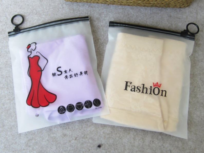 Pu&ograve; includere: Due sacchetti di plastica trasparente con cerniera e cerniere nere. Un sacchetto contiene un tessuto viola con una figura femminile rossa che indossa un vestito rosso e il testo "S 女人  秀出好身材". L'altro sacchetto contiene un tessuto beige con il testo "Fashion" e una corona.