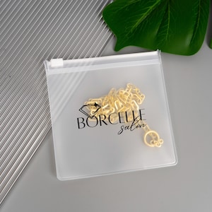 May include: A gold-colored necklace inside a clear plastic zip bag. The bag has the text "BORCELLE salon" printed on it in black. The background is a light gray surface with a green leaf in the upper right corner.