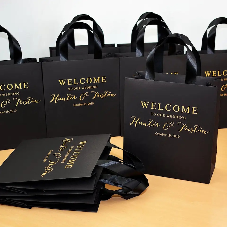 May include: Black gift bags with gold lettering and black ribbon handles. The bags read "WELCOME TO OUR WEDDING" followed by names and a date. Several bags are visible, some stacked, others upright.