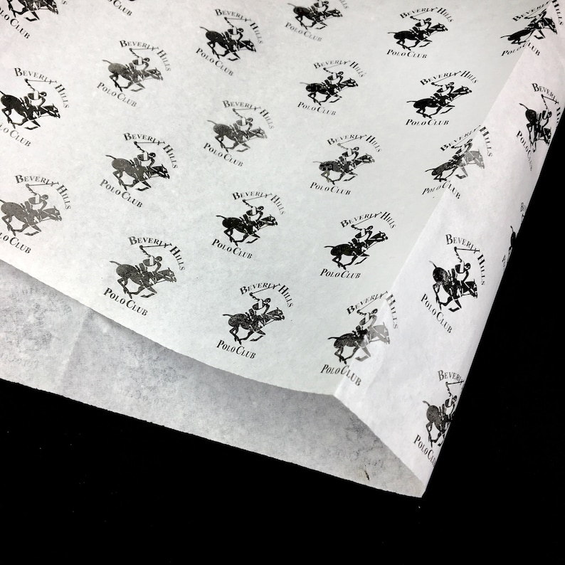 K&ouml;nnte beinhalten: Wei&szlig;es Seidenpapier mit einem sich wiederholenden schwarzen Muster eines Polospielers auf einem Pferd, mit dem Text "Beverly Hills Polo Club". Das Papier ist gefaltet und zeigt seine d&uuml;nne Textur und den schwarzen Hintergrund.