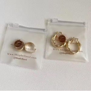 Può includere: Due paia di orecchini color oro in sacchetti di plastica trasparente. Un paio è un piccolo design rotondo, l'altro è uno stile a cerchio intrecciato. Le borse hanno un logo e un indirizzo web stampati.