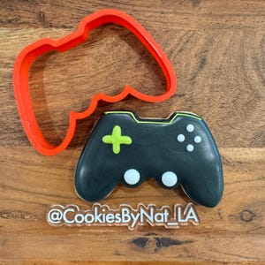 Może przedstawiać: Ciasteczko w kształcie czarnego kontrolera do gier z zielonymi i białymi akcentami, obok czerwonej plastikowej foremki do ciastek w kształcie kontrolera. Ciasteczko ma zielony krzyż i białe przyciski. Widoczny jest tekst "@CookiesByNat_LA".