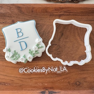 May include: A decorated sugar cookie in the shape of a shield with the letters "B" and "D" in blue icing. The cookie is adorned with white roses and green leaves. A white plastic cookie cutter is next to the cookie. The text "@CookiesByNat_LA" is at the bottom.