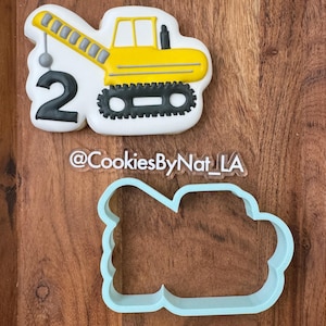 May include: A yellow construction vehicle cookie cutter with a ball on a chain, a number 2, and the text "@CookiesByNat_LA".