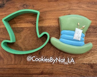 May include: A green Mad Hatter hat cookie cutter next to a decorated Mad Hatter hat cookie. The cookie is green with blue stripes and a tag that reads "10/6". The cookie cutter and cookie are on a wooden surface.