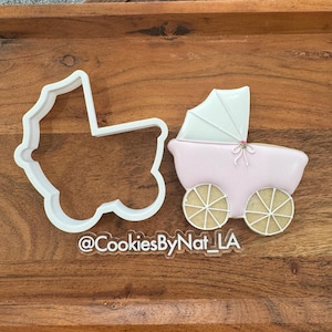 May include: A pink baby carriage cookie decorated with white icing and a white sunshade. Next to it is a white plastic cookie cutter in the shape of a baby carriage. The text "@CookiesByNat_LA" is visible.