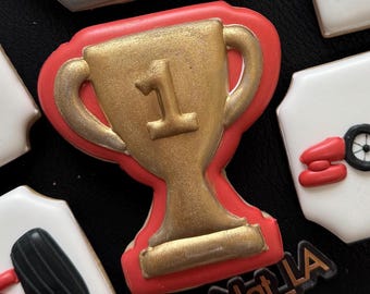 May include: A red and gold trophy cookie with the number "1" on the front. The cookie is decorated with a gold metallic finish and has a red outline. The cookie is on a black background.