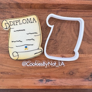 May include: A diploma-shaped cookie with the word "DIPLOMA" on it, next to a white plastic cookie cutter. The cookie is decorated with black lines and a blue and yellow seal. The cookie is on a wooden surface.