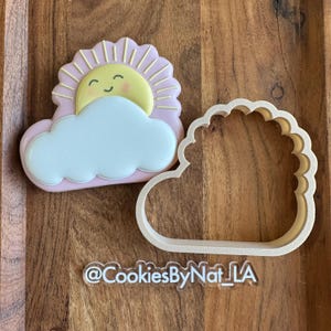 May include: A pink and yellow cookie cutter in the shape of a smiling sun behind a cloud. The cutter is on a wooden surface with the text "@CookiesByNat_LA" below it.
