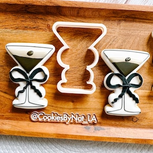 May include: Two decorated sugar cookies in the shape of a martini glass with a black bow and a white and green filling. A white plastic cookie cutter in the shape of a martini glass is in the center of the image. The text "@CookiesByNat_LA" is on the wooden surface below the cookies.