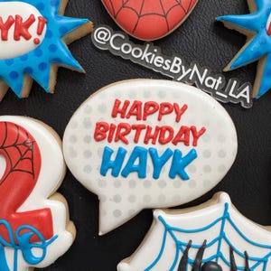 May include: A set of five decorated sugar cookies featuring a red and blue superhero theme. The cookies are decorated with red, blue, and white icing. One cookie has a speech bubble with the text "Happy Birthday Hayk".