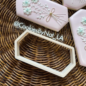 May include: A white plastic cookie cutter shaped like a hexagon with rounded corners. The cutter is on a woven brown wicker surface. The text "@CookiesByNat_LA" is visible in the background.