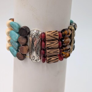 May include: A stack of beaded bracelets in various colors and textures. The bracelets include turquoise, brown, silver, red, and amber beads. The bracelets are displayed on a white cylindrical object.