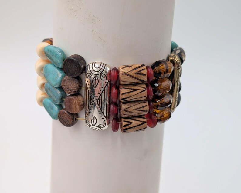 May include: A stack of beaded bracelets in various colors and textures. The bracelets include turquoise, brown, silver, red, and amber beads. The bracelets are displayed on a white cylindrical object.