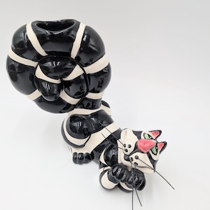 May include: A whimsical ceramic cat figurine in black and white stripes. The cat has pink accents on its nose and ears, with green eyes and black whiskers. The figurine has a unique, stylized design.