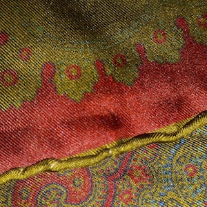 May include: Close-up of a fabric with a paisley pattern. The fabric is red, green, and blue. The fabric is folded over, showing the underside of the fabric, which is a solid gold color.