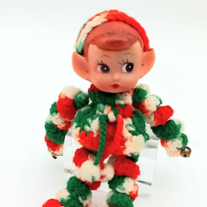 May include: A vintage Christmas elf doll with a plastic face and red hair, wearing a crocheted outfit in red, green, and white. The doll has bells on its feet and hands.