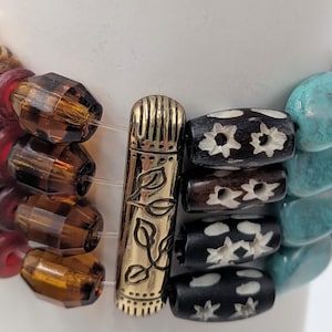May include: A multi-strand bracelet featuring a variety of beads. The bracelet includes wooden beads with geometric patterns, faceted amber-colored beads, and turquoise-colored stone beads. A gold-colored metal clasp with leaf details secures the bracelet.