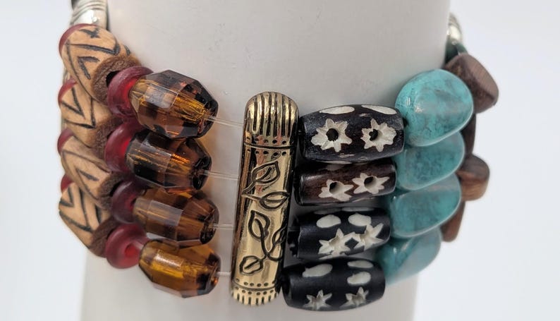 May include: A multi-strand bracelet featuring a variety of beads. The bracelet includes wooden beads with geometric patterns, faceted amber-colored beads, and turquoise-colored stone beads. A gold-colored metal clasp with leaf details secures the bracelet.