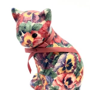 May include: A ceramic cat figurine with a floral pattern of red, purple, yellow, and green pansies. The cat has a pink ribbon tied around its neck. The figurine is sitting upright with its head turned slightly.