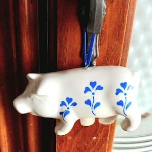 May include: A white ceramic pig ornament with blue floral designs. The pig has a blue ribbon for hanging. The ornament is hanging on a wooden surface.