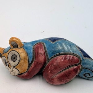 May include: A ceramic figurine of a stylized cat in a reclining pose. The cat features a blue body with red paws and a tan face with large, round eyes. The ears are tan, and the design includes dark blue geometric shapes.