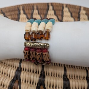 May include: Four stacked bracelets with a variety of beads. The top bracelet has turquoise, cream, and brown beads. The second bracelet has amber-colored faceted beads. The third bracelet has a gold-colored metal accent with red beads. The bottom bracelet has wooden beads.