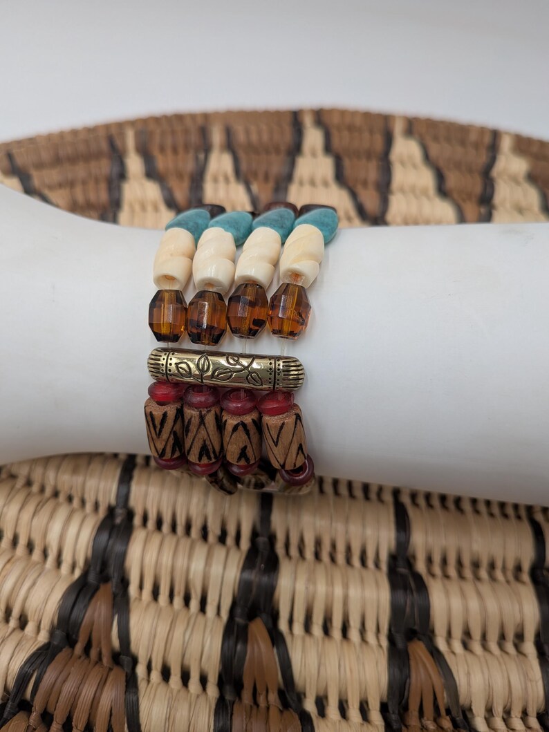 May include: Four stacked bracelets with a variety of beads. The top bracelet has turquoise, cream, and brown beads. The second bracelet has amber-colored faceted beads. The third bracelet has a gold-colored metal accent with red beads. The bottom bracelet has wooden beads.