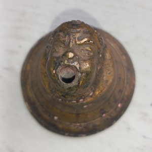 May include: A vintage brass spout with a face sculpted on the front. The spout has a round opening and is attached to a larger, round base.
