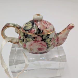 May include: A miniature porcelain teapot with a floral rose pattern in pink, white, and green. The teapot has gold trim and a small white ribbon attached. The teapot is resting on a clear, faceted display stand.
