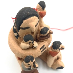 May include: A hand-crafted clay figurine of a Native American woman holding four children. The woman is wearing a traditional dress with white dots. The children are all wearing simple clothing. The figurine is painted in brown, black, and red.