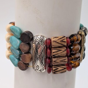 May include: A multi-strand bracelet with a variety of beads. The bracelet features turquoise, cream, and brown beads, along with wooden and red beads. Silver-toned metal accents and amber-colored beads complete the design. The bracelet is displayed on a white cylindrical object.