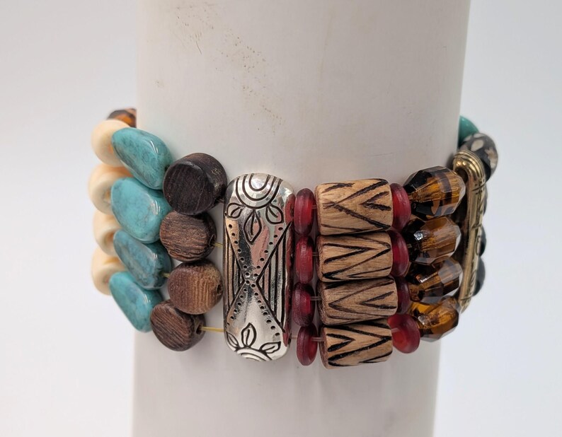May include: A multi-strand bracelet with a variety of beads. The bracelet features turquoise, cream, and brown beads, along with wooden and red beads. Silver-toned metal accents and amber-colored beads complete the design. The bracelet is displayed on a white cylindrical object.