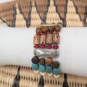 May include: A multi-strand bracelet with a variety of beads in brown, red, silver, turquoise, and cream colors. The bracelet features wooden, metal, and glass beads, creating a layered and textured look. The bracelet is displayed on a white surface.