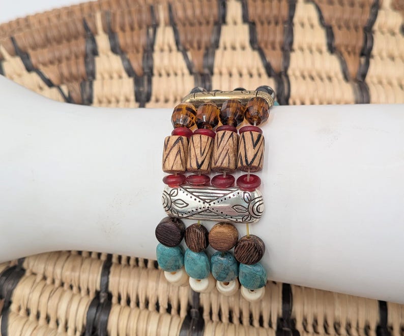 May include: A multi-strand bracelet with a variety of beads in brown, red, silver, turquoise, and cream colors. The bracelet features wooden, metal, and glass beads, creating a layered and textured look. The bracelet is displayed on a white surface.