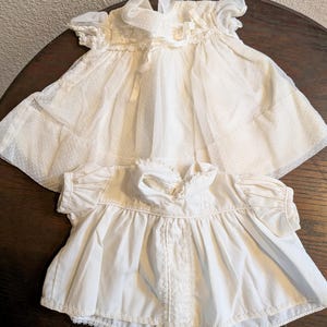May include: Two vintage white baby dresses. The top dress has a sheer overlay with a polka dot pattern and ruffled sleeves. The bottom dress is a solid white with lace trim and short sleeves. Both dresses are on a dark brown surface.