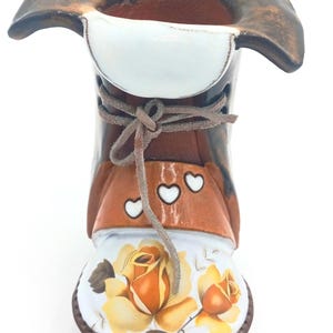 May include: A ceramic boot-shaped vase with a brown and white glaze. The vase is decorated with a floral design featuring yellow roses and white hearts. The vase has a brown leather-like lace tied around the top.