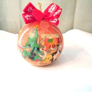 May include: A round Christmas ornament featuring Bugs Bunny, Daffy Duck, Sylvester the Cat, and Tweety Bird. The ornament is decorated with a red bow and the words "Looney Tunes".